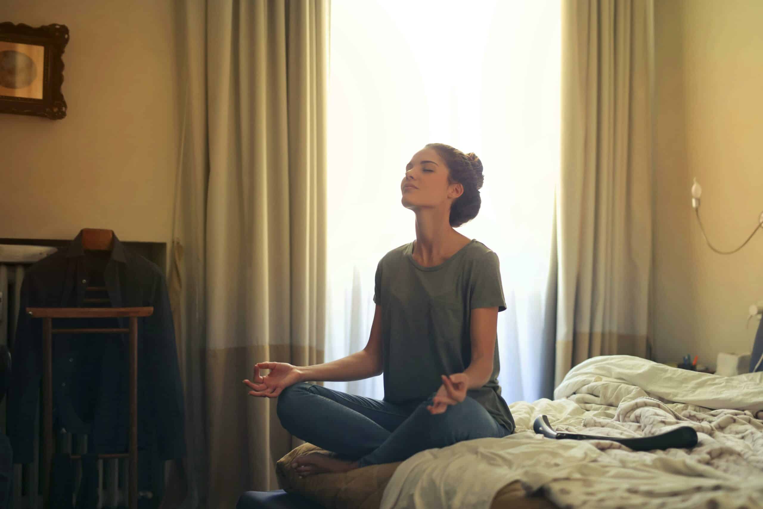 Adult woman practicing meditation on her bed surrounded by a calm bedroom atmosphere.