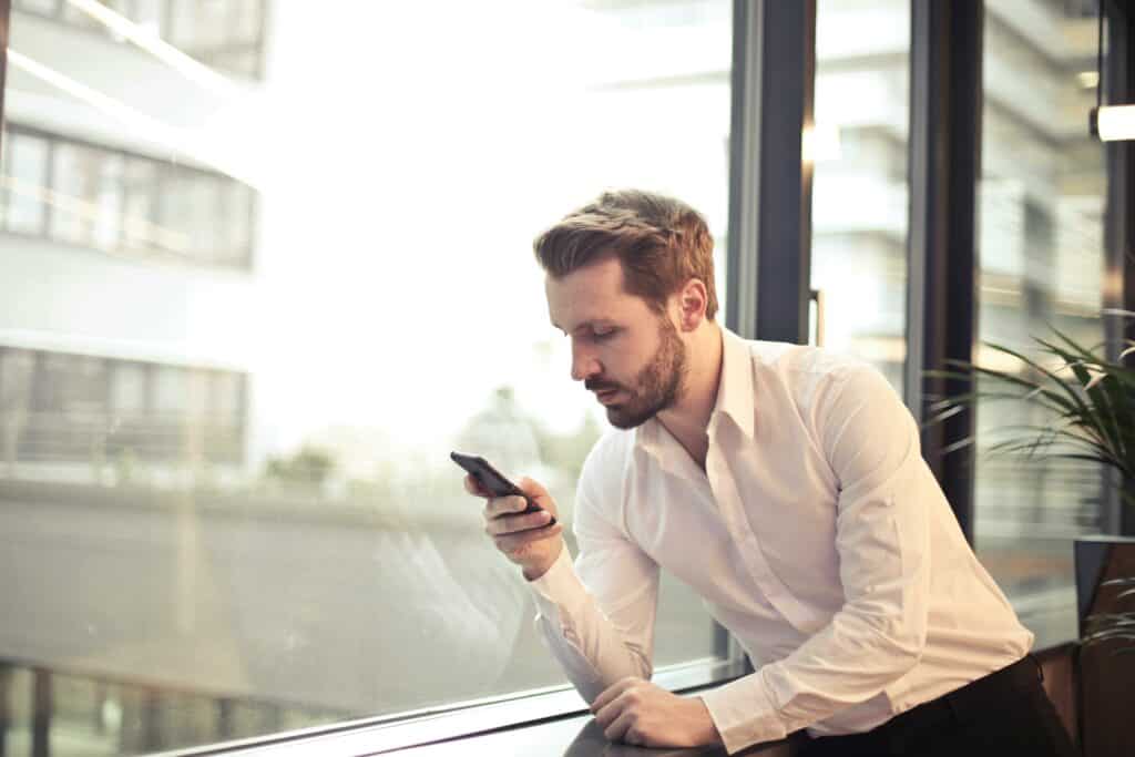 A businessman in a white shirt texts on his smartphone by a large glass window inside an office.